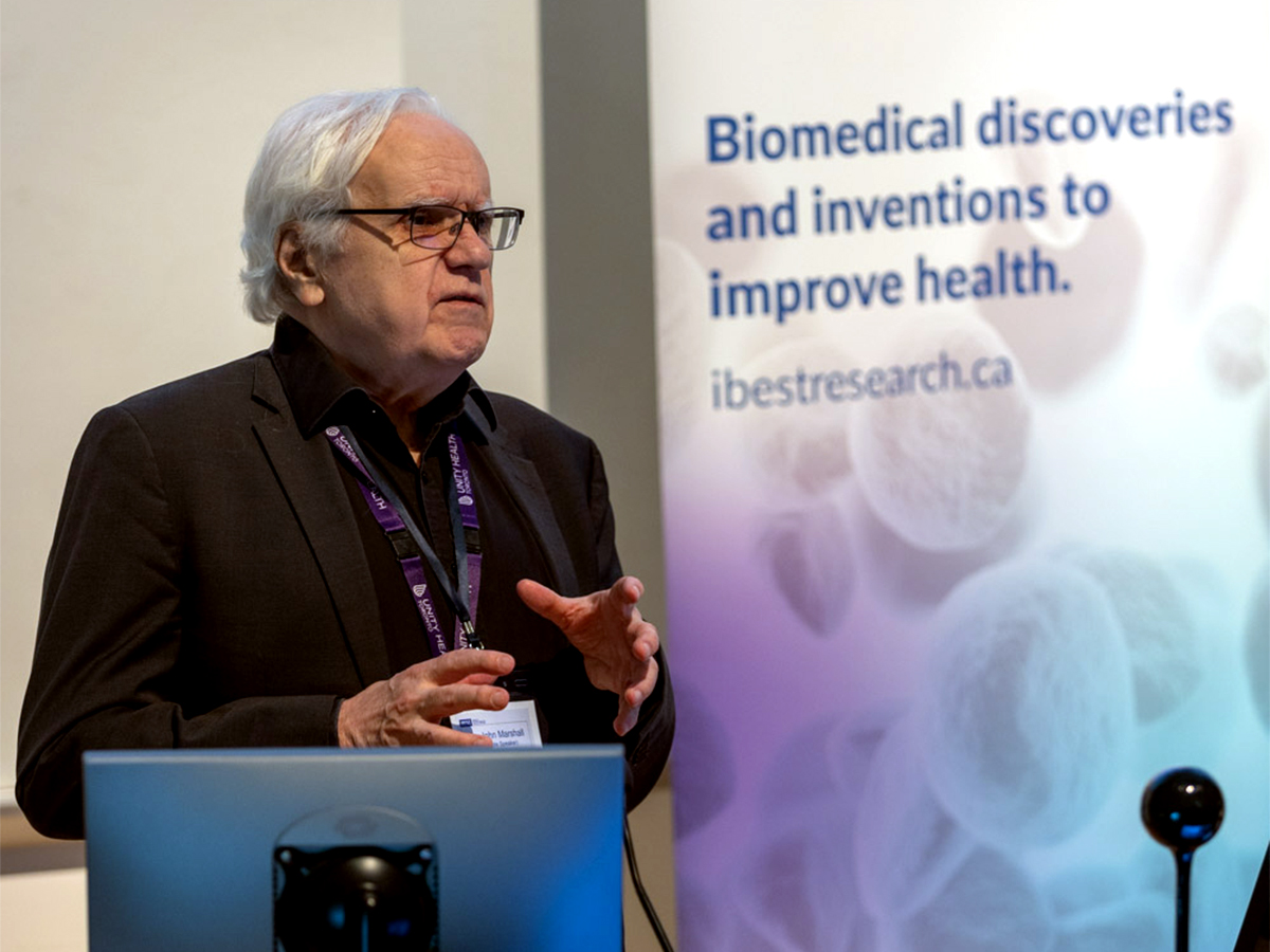 Dr. John Marshall, wearing glasses and a dark suit jacket, in front of an iBEST banner. 